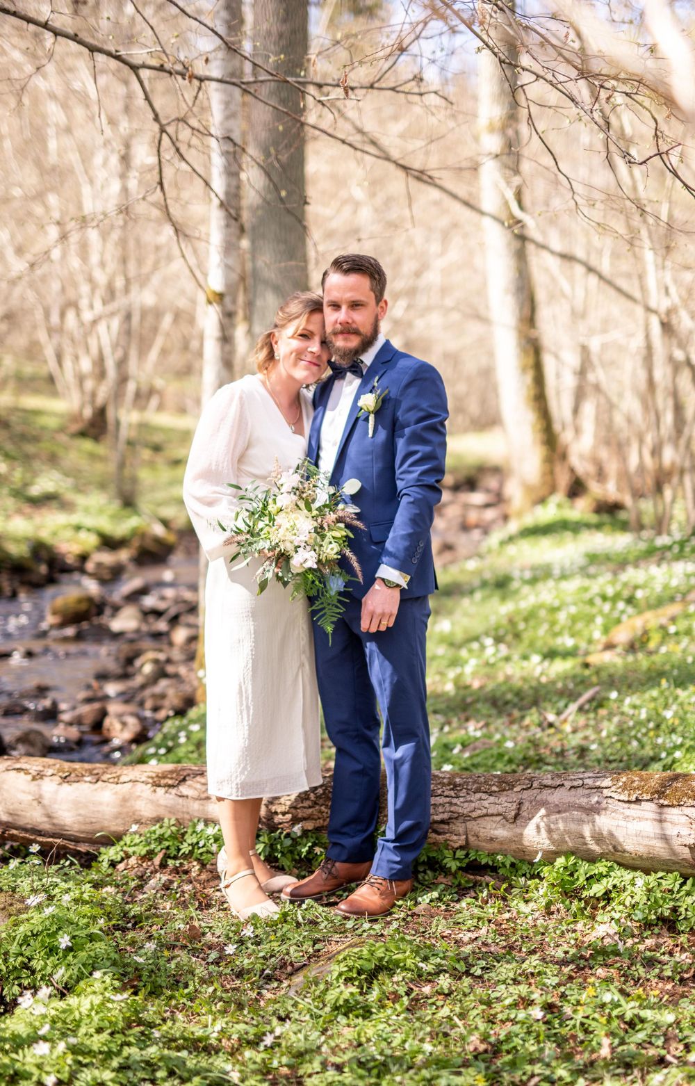 Bride and groom pose together in a sunlit forest holding a white bouquet on their wedding day.