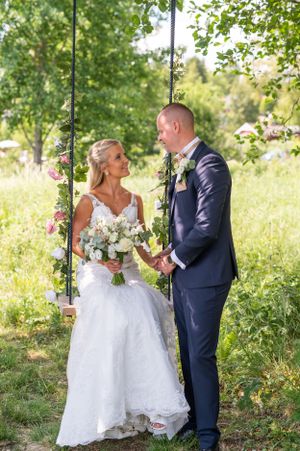 Bride sitting on a floral decorated swing holding bouquet while groom stands beside her outdoors.
