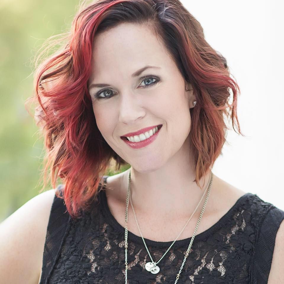 Smiling woman with red-highlighted hair wearing black lace top and silver necklace outdoors.
