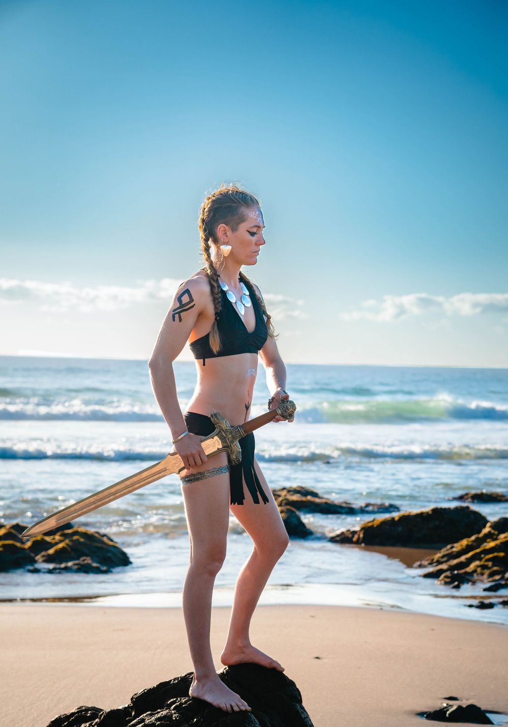 Warrior-styled figure poses with sword on beach rocks at sunset.