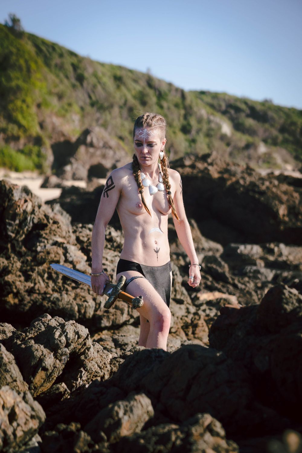 Athletic topless woman traverses rocky coastal terrain holding a viking sword.