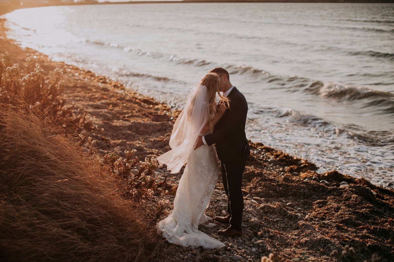 Couple in wedding attire embracing on a beach at sunset.