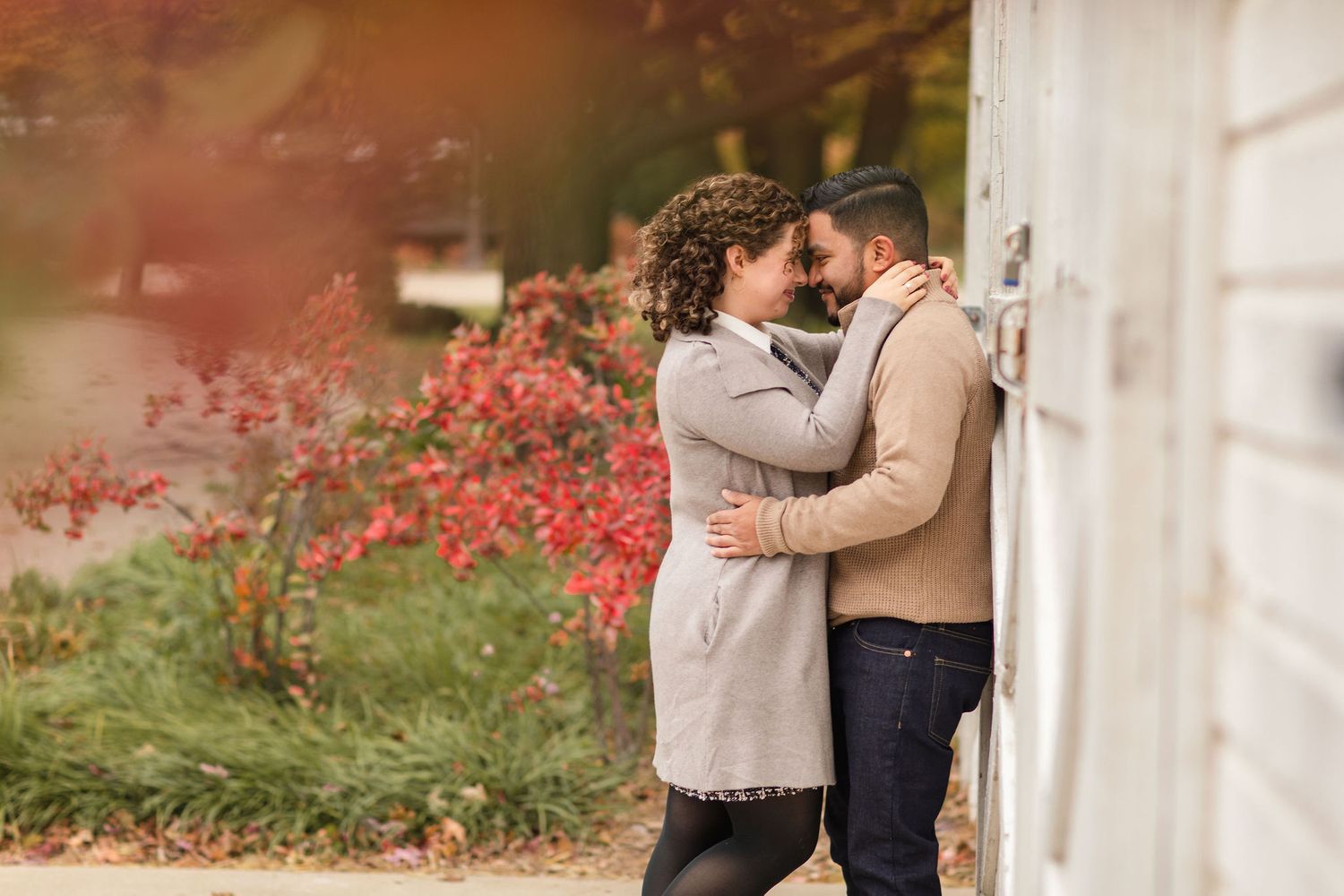 Romantic autumn moment between couple near white building with red foliage in background.
