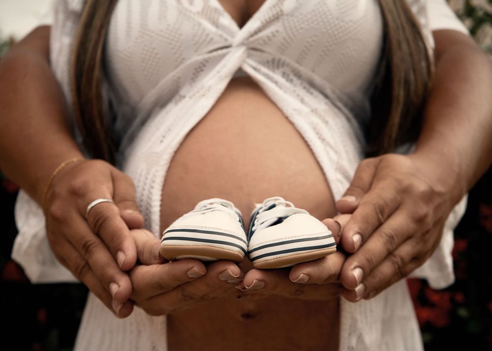 Small baby shoes held against a pregnant belly in a white dress during a maternity photoshoot.