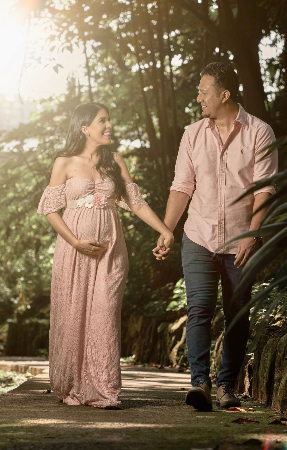 A couple walks hand in hand on a sunlit forest path during a maternity photoshoot.