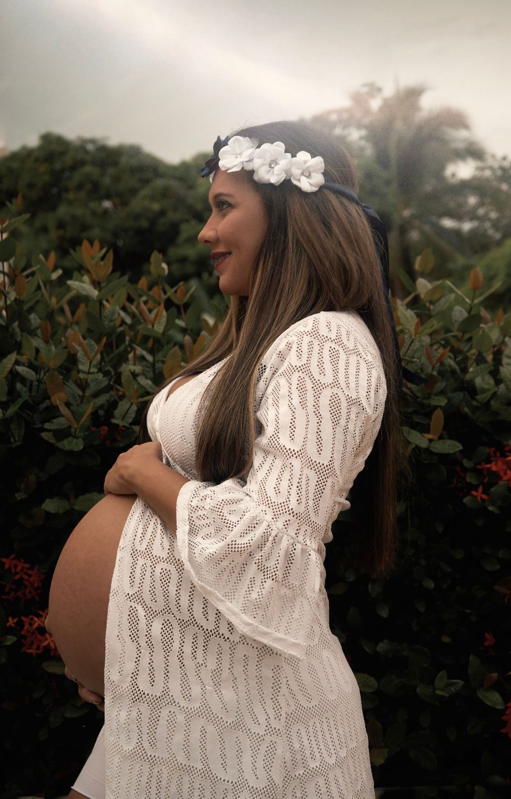Expectant woman in white lace dress and floral crown poses in garden setting.