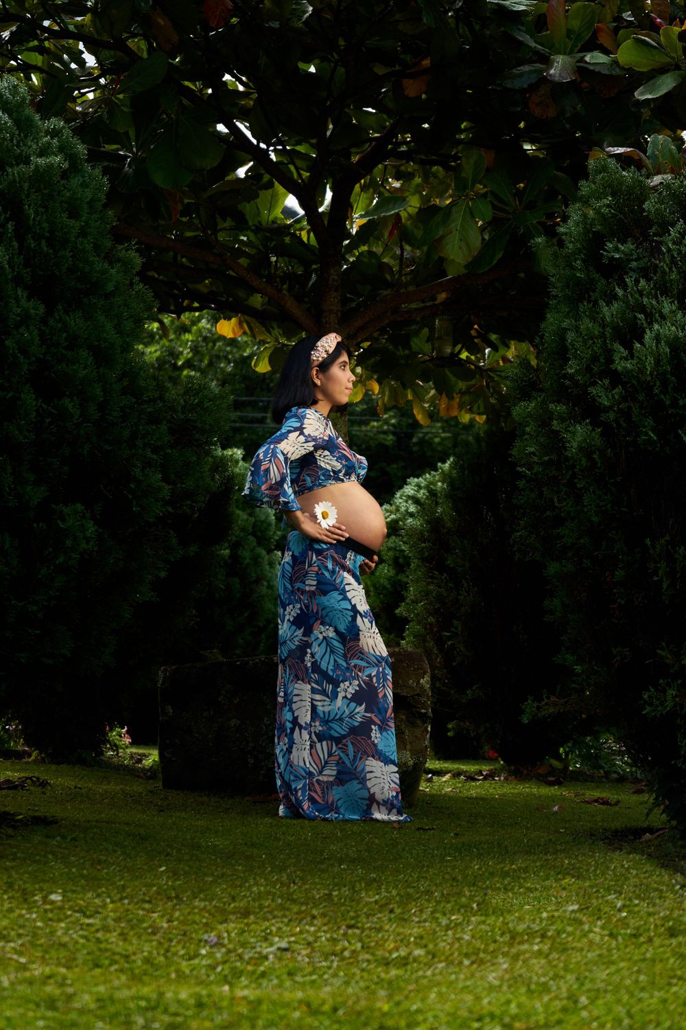 A woman in a blue floral dress stands in a lush garden setting showing her pregnant belly.