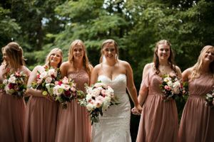 Bridesmaids in dusty rose dresses laugh while holding pink and white bouquets.
