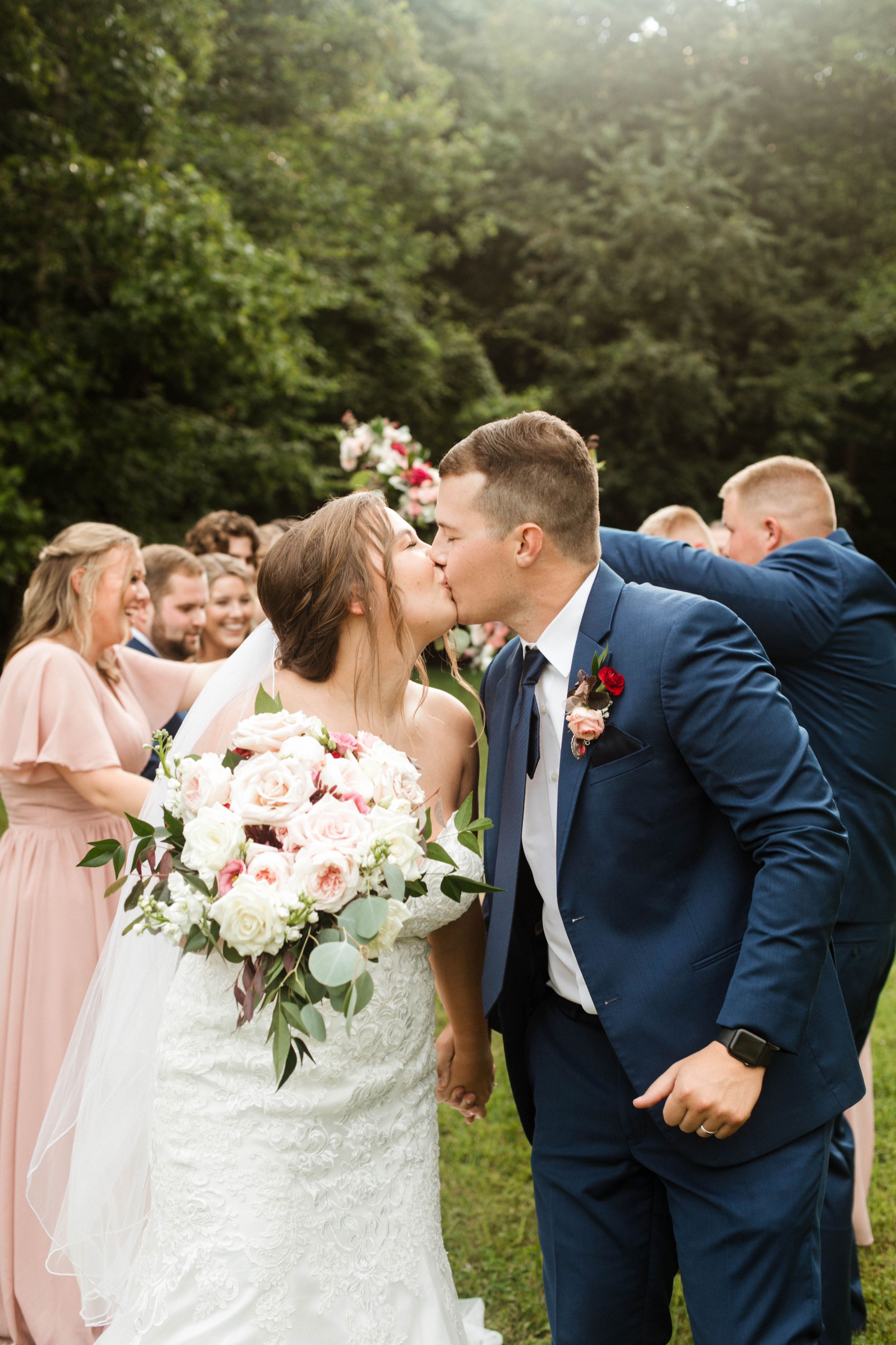 A wedding party celebrates outdoors as the newlyweds share a kiss.