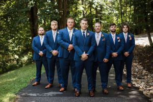 Groomsmen in matching navy suits and brown shoes pose together on a wooded path.