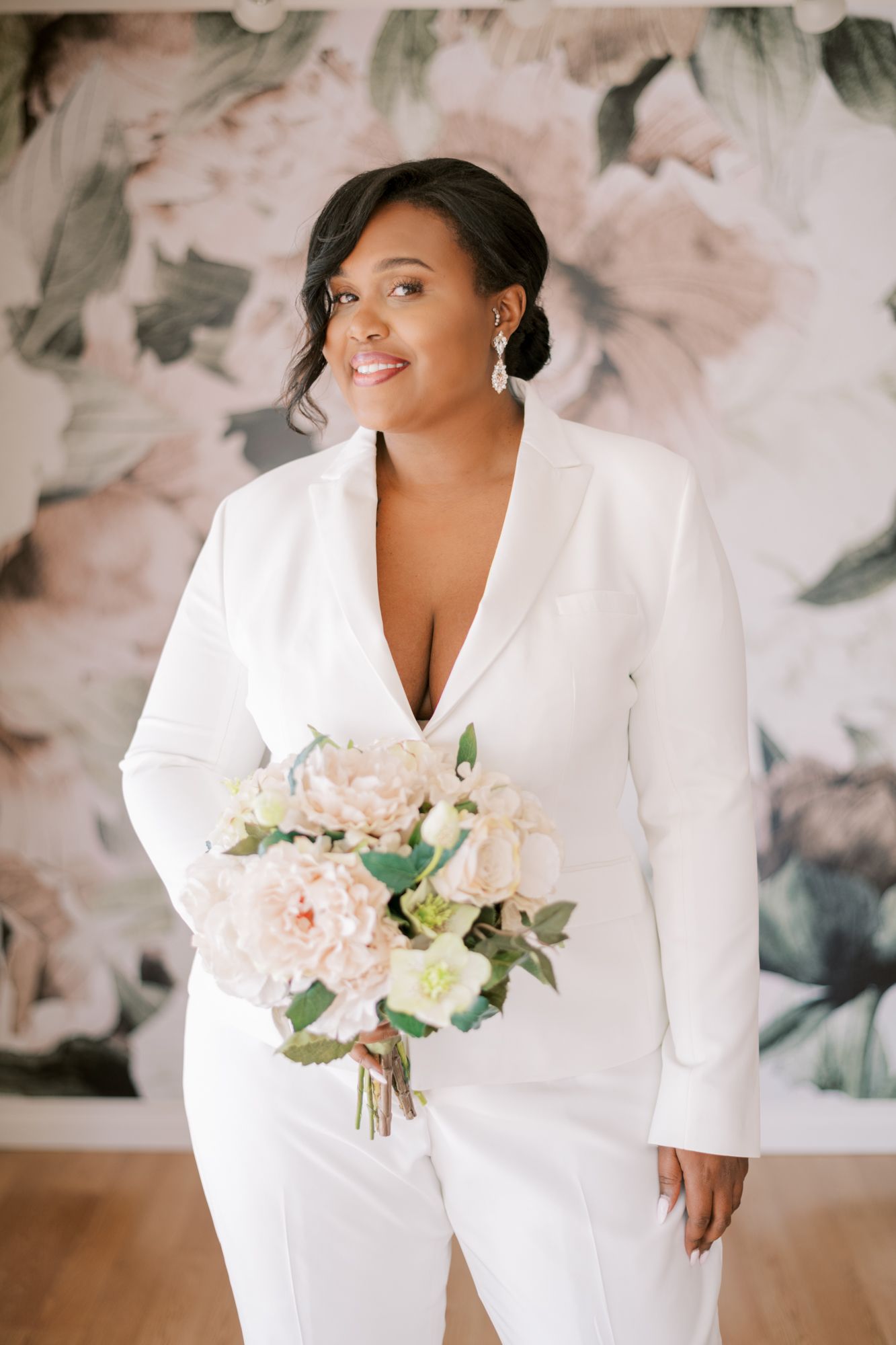 Elegant bride in white pantsuit holding soft pink floral bouquet against abstract backdrop.