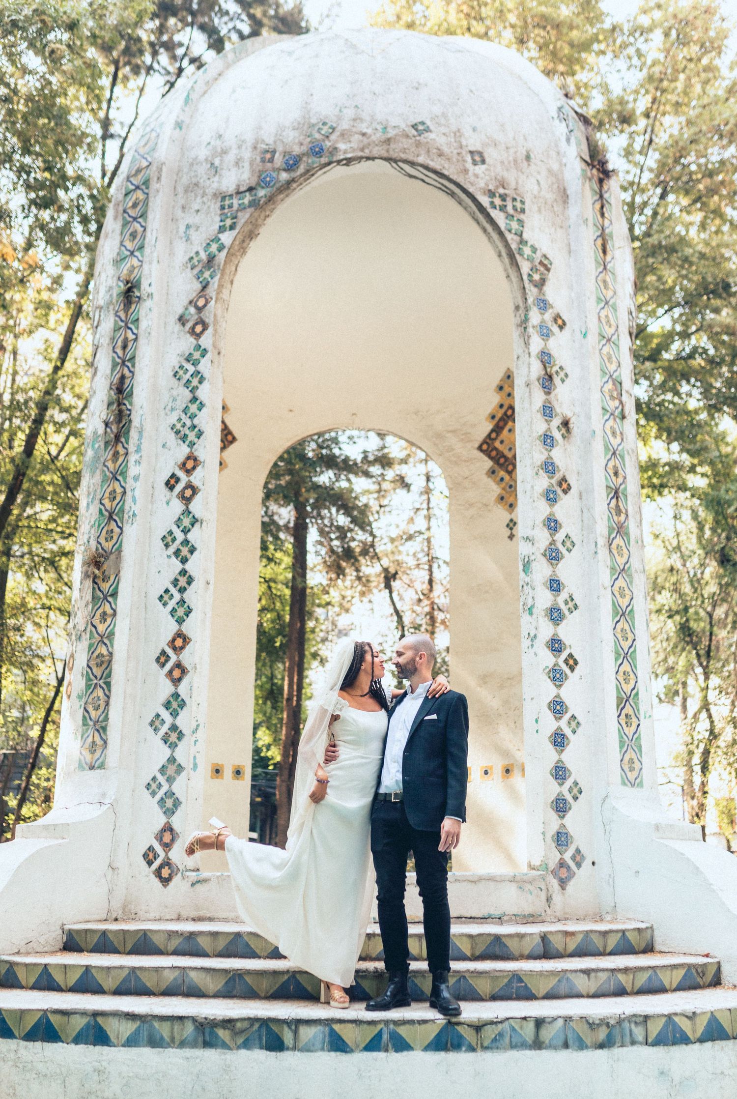 Newlyweds standing under ornate white archway decorated with geometric tiles at outdoor venue.