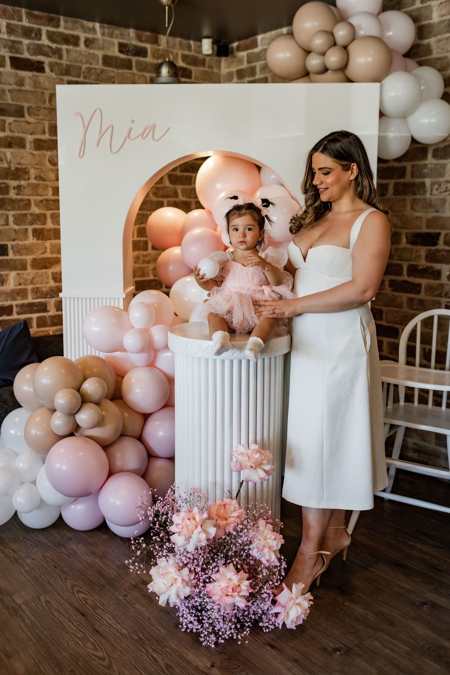 Elegant party setup with pink balloons and floral arrangements against a rustic brick wall backdrop.