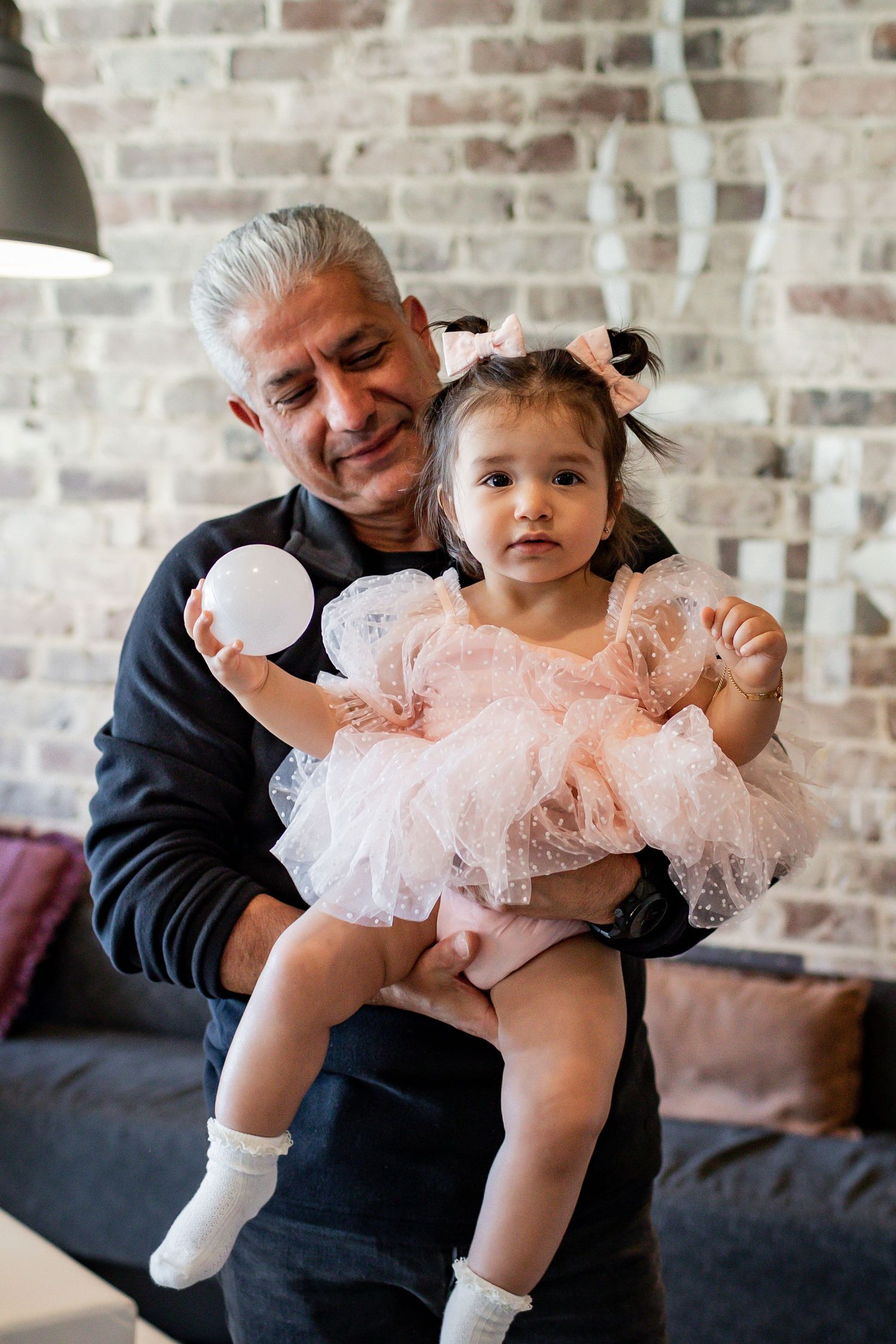 Child in pink dress sitting with adult at celebration with brick wall backdrop.