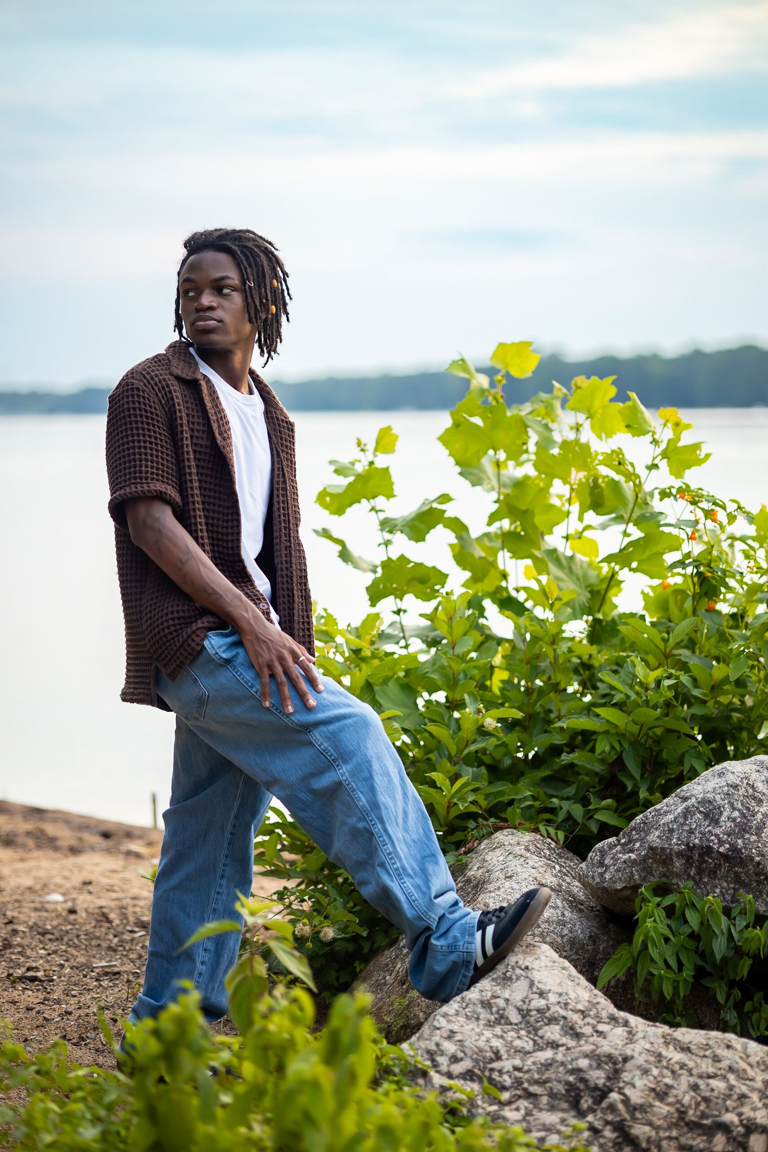 Young man in brown shirt and jeans posing against waterfront foliage.
