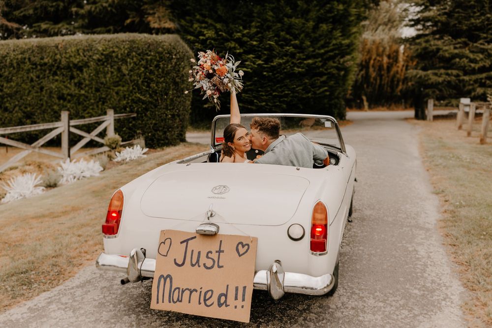 Classic white convertible decorated with 'Just Married' sign and flowers driving down a tree-lined path.