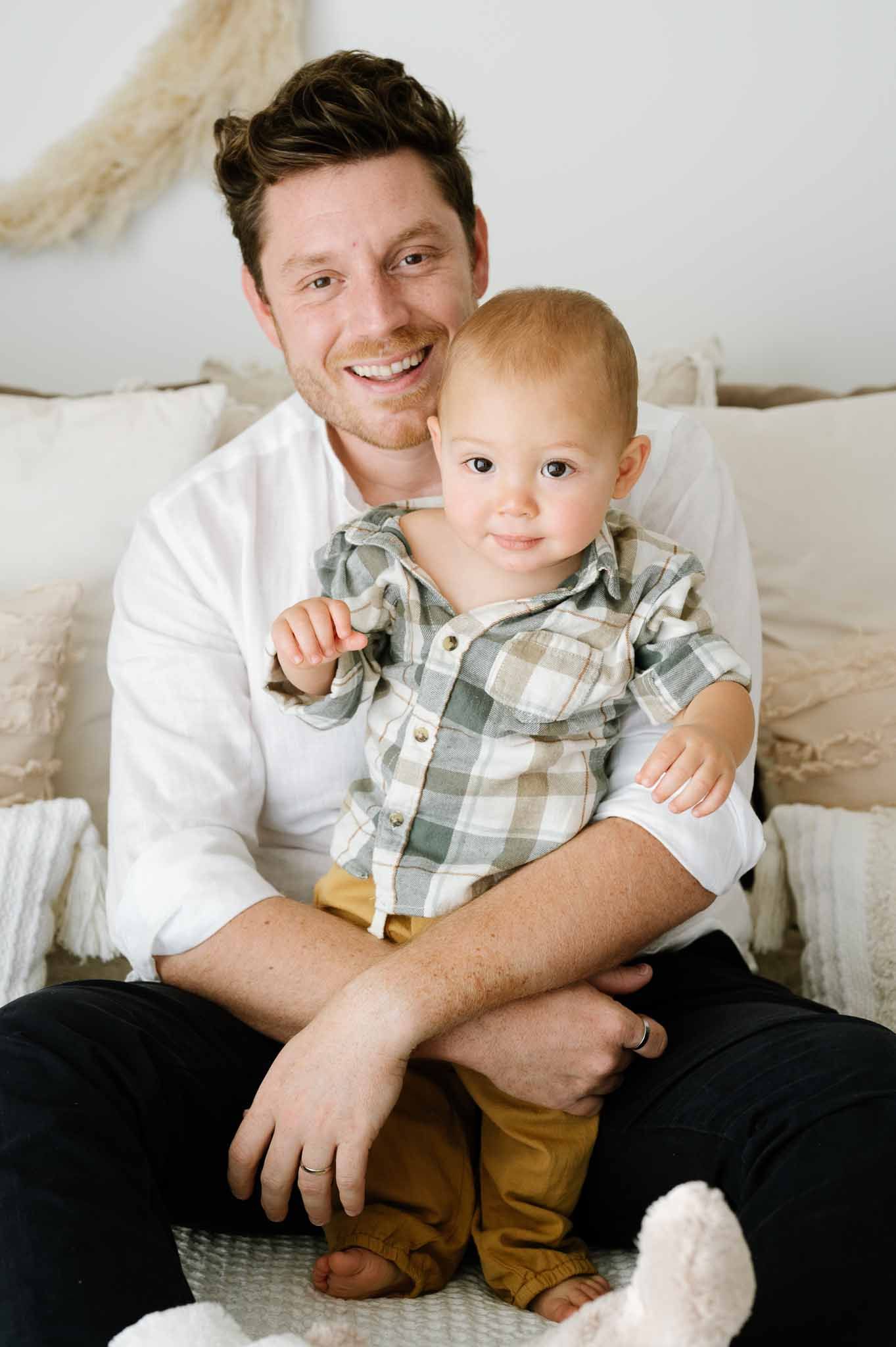 Parent and toddler in plaid shirt sitting together on white couch with pampas grass decor.