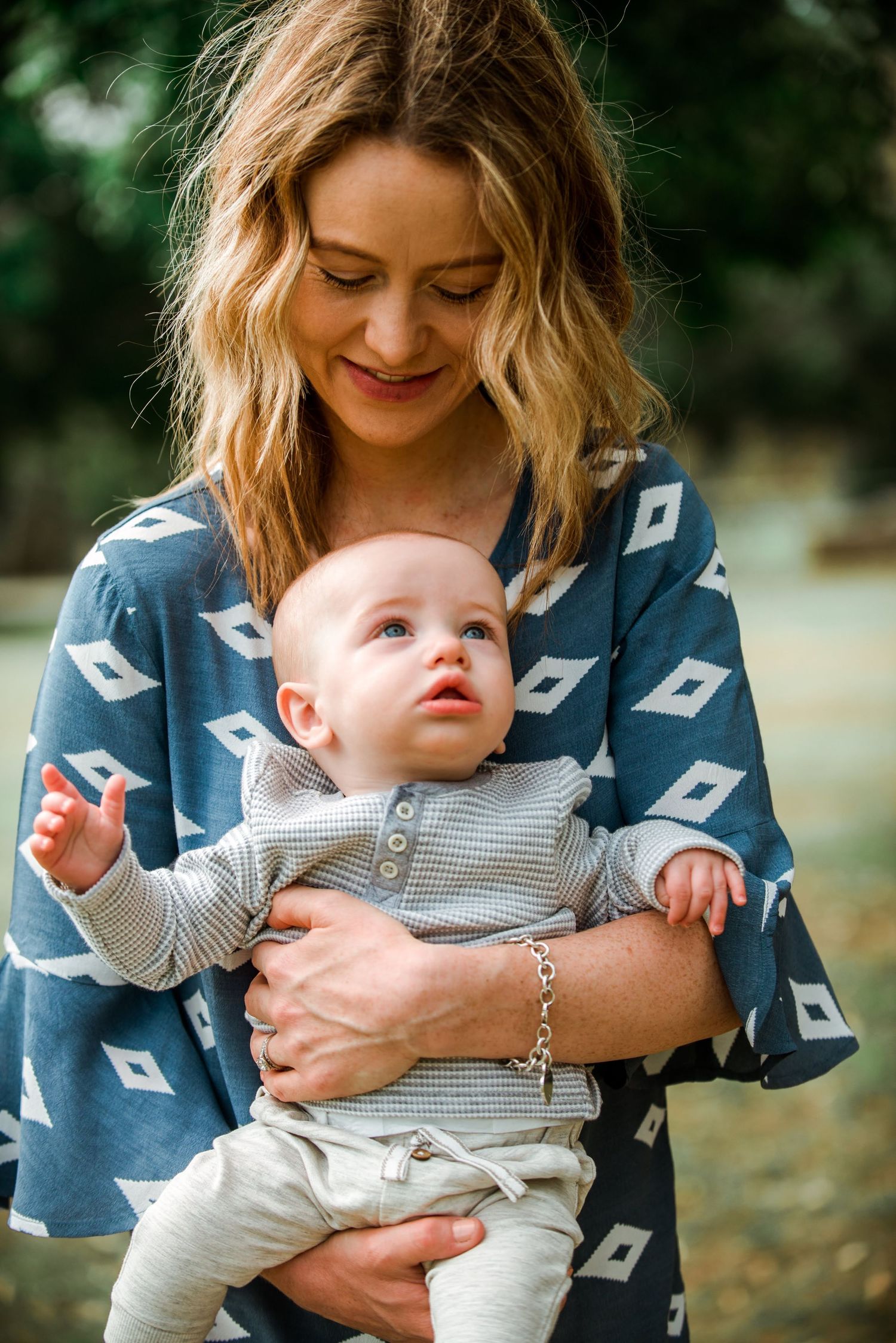 Woman in patterned blue dress holding smiling baby wearing grey outfit against blurred green backdrop.