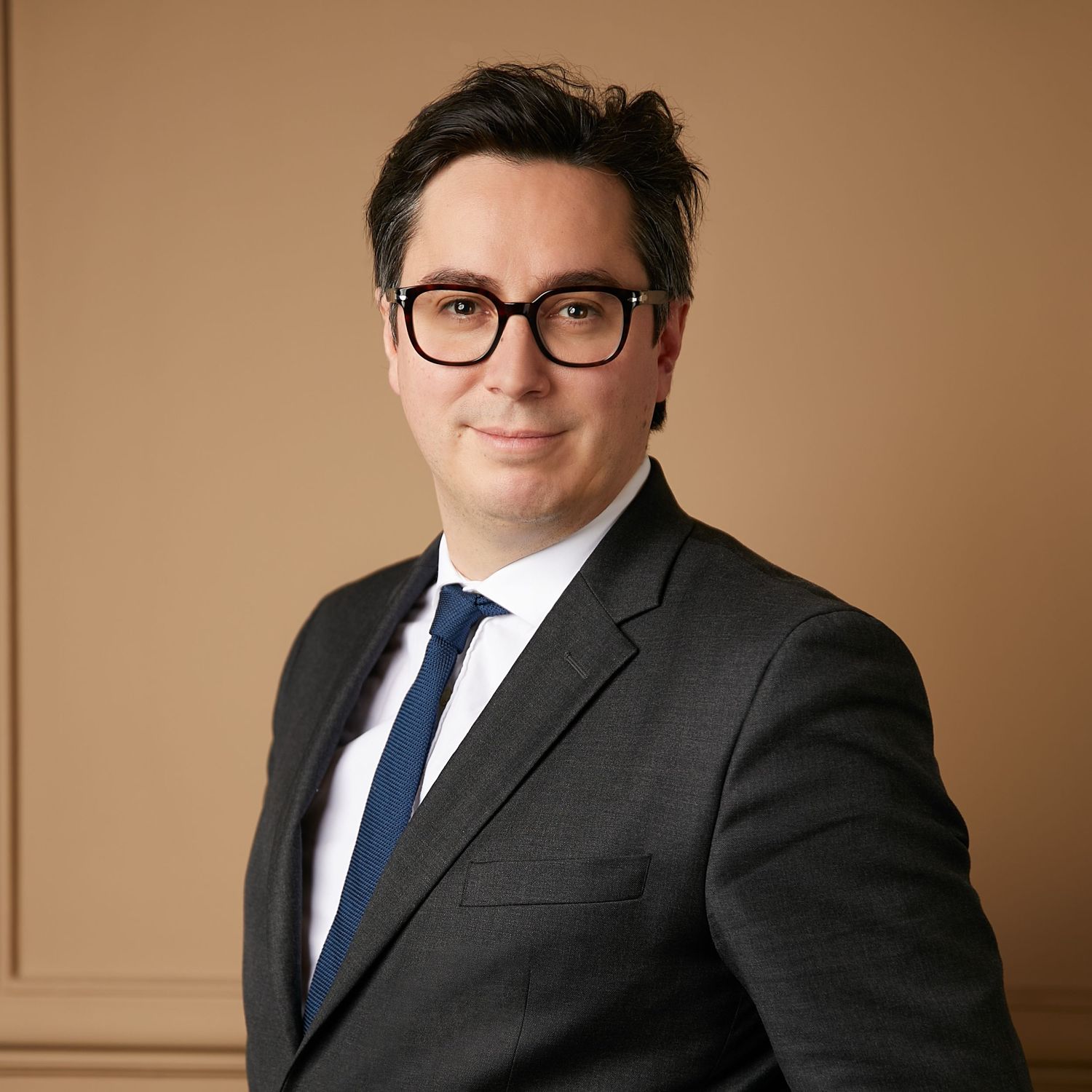 Professional headshot of a person wearing glasses, dark suit and blue tie against neutral backdrop.