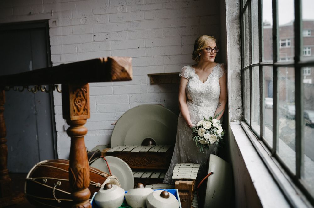 Natural light wedding photo with classic lace gown and bouquet by industrial window.