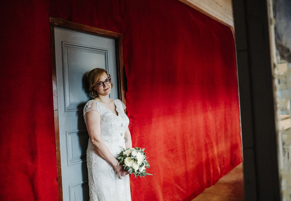 Dramatic bridal portrait against vibrant red wall featuring lace wedding dress and bouquet.