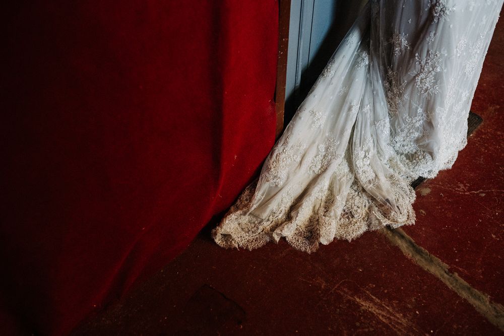 Detail shot of lace wedding dress hem against red velvet backdrop and wooden floor.