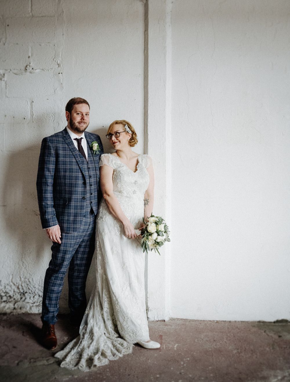 Full-length wedding portrait featuring plaid suit and lace gown against white industrial wall.