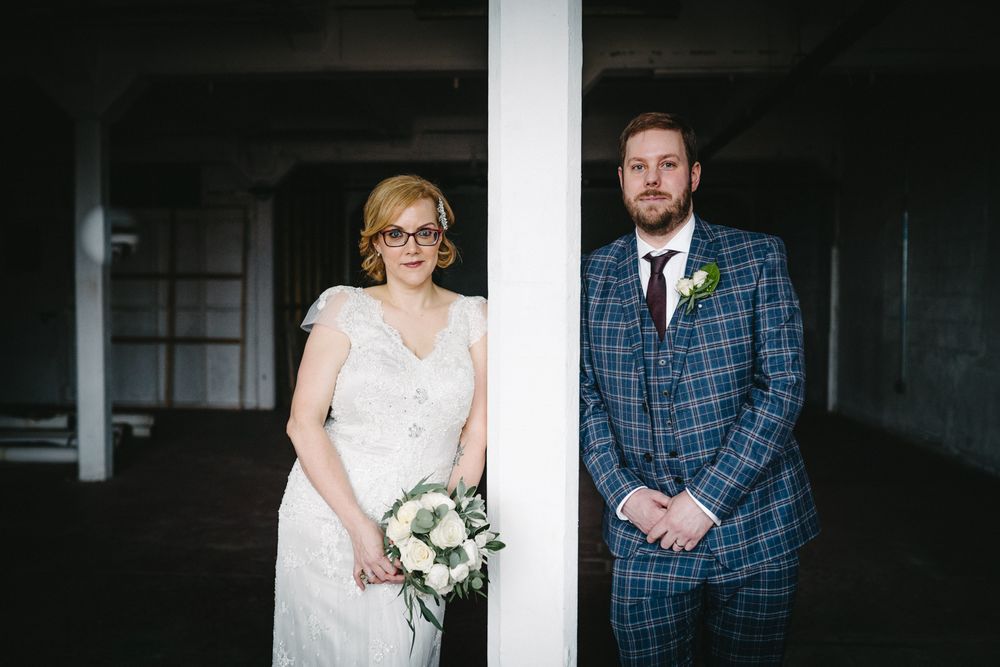 A bride and groom stand separated by a white column in a moody industrial setting.