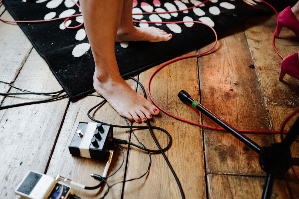 Bare feet near audio equipment and cables on a wooden floor with a black patterned rug.