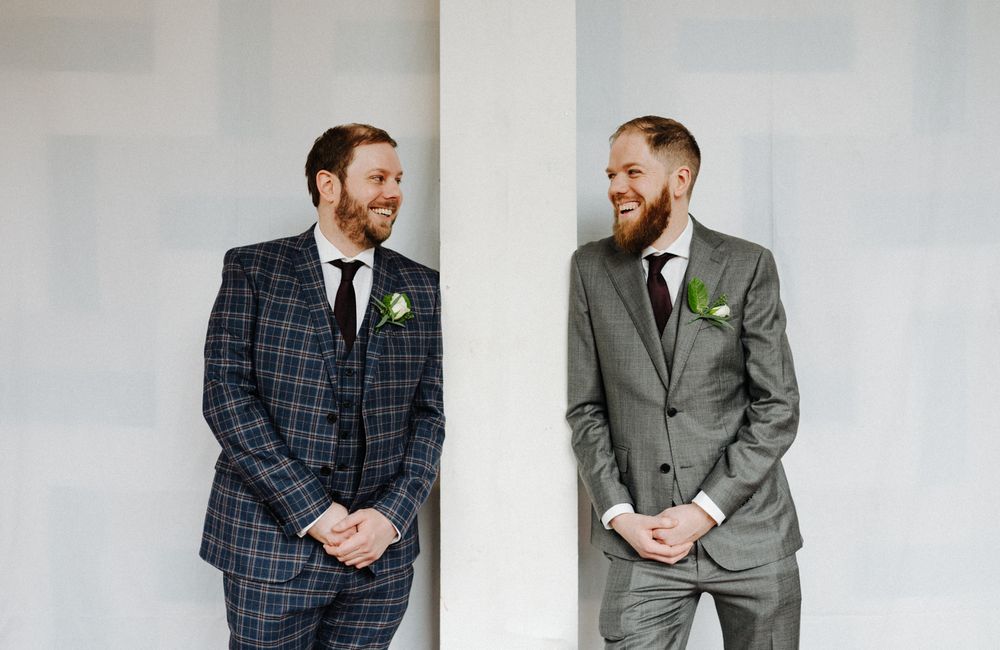 Two men wearing formal suits share a moment of laughter against a white wall.