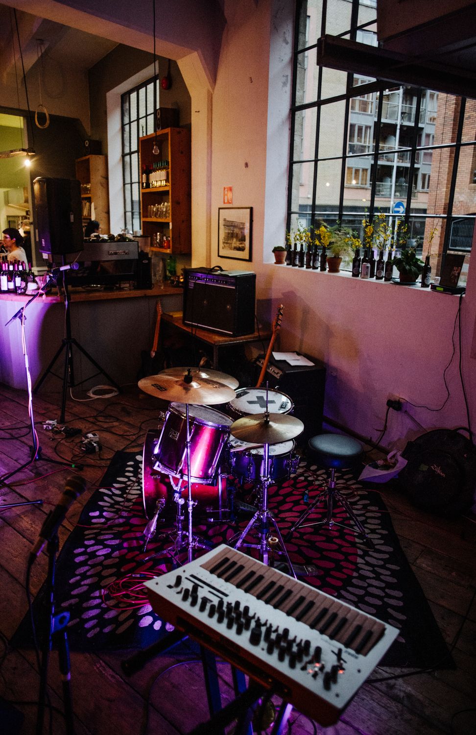 Music stage setup with drum kit, keyboard and equipment in front of large windows.