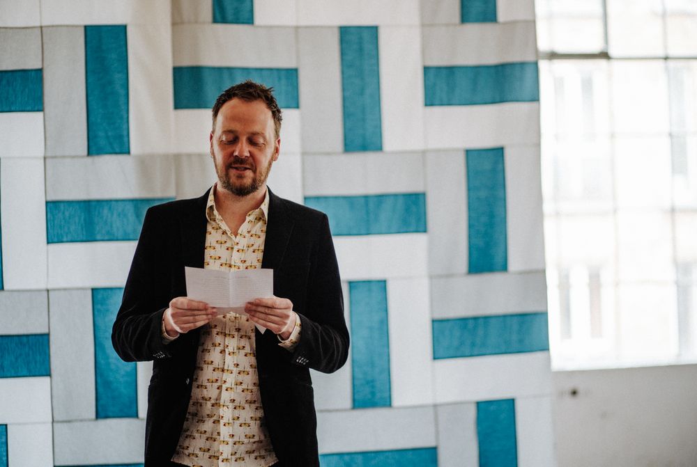 Speaker stands against geometric turquoise wall pattern reading from papers.