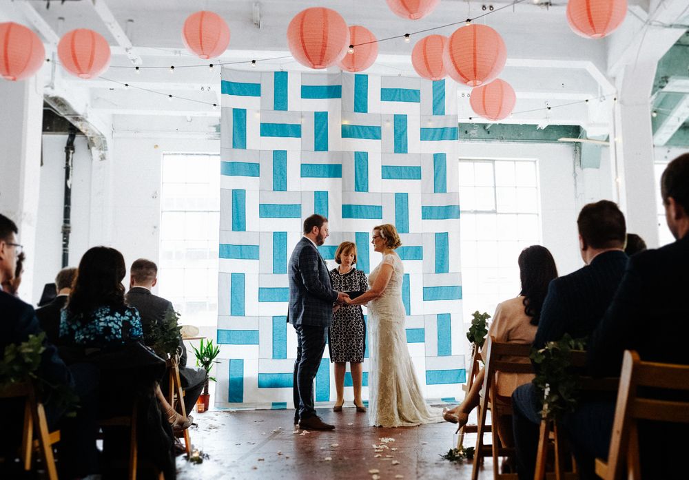 Modern wedding ceremony with geometric blue backdrop and coral paper lanterns in an industrial venue.