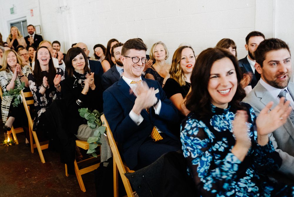 Wedding guests clapping and smiling during a reception celebration in an industrial space.