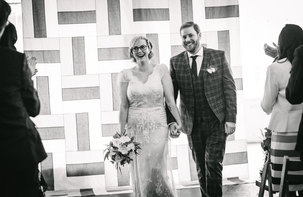 Black and white photo of newlyweds walking past geometric wall art at modern venue.