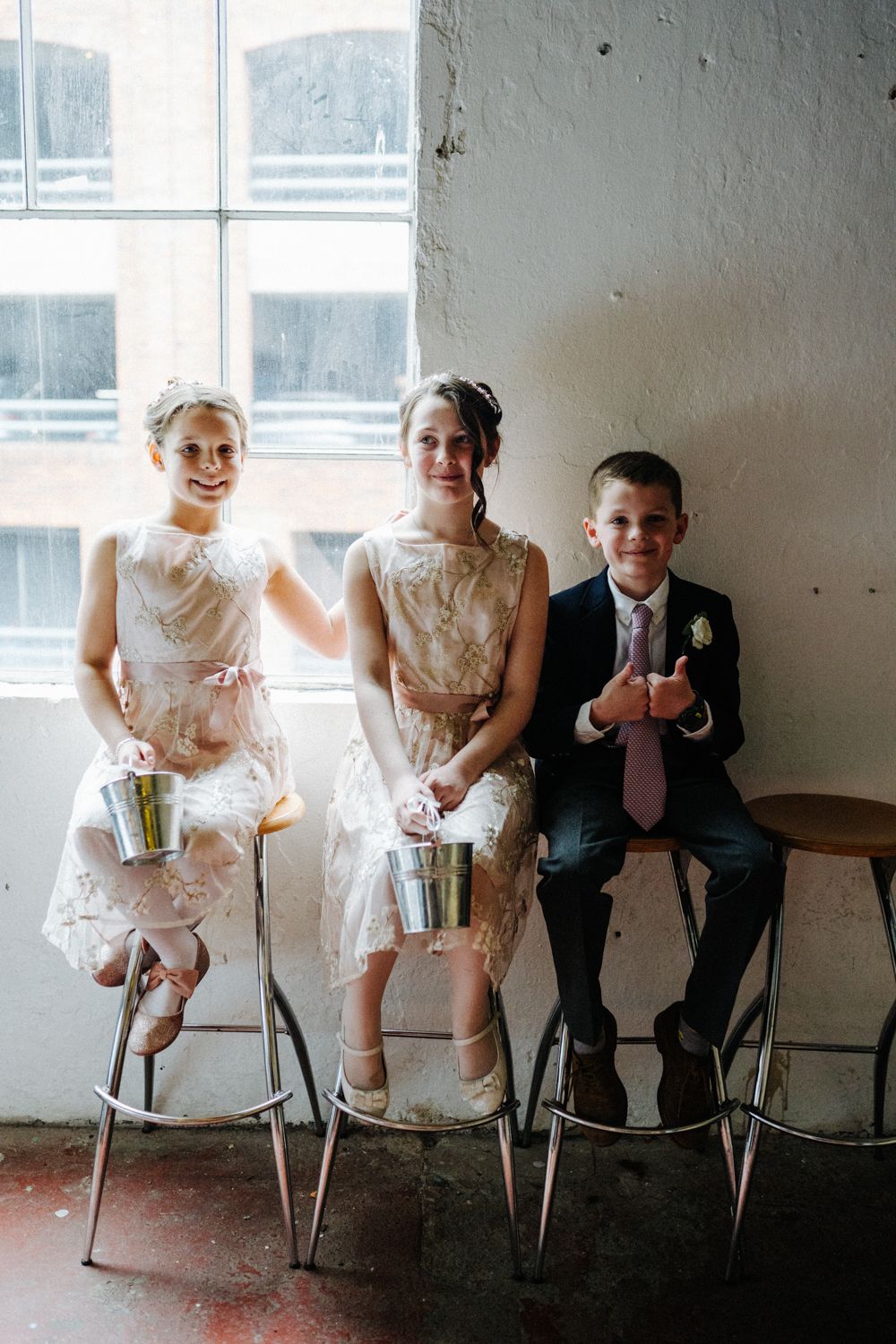 Three children in formal attire sitting on barstools by a window in vintage setting.
