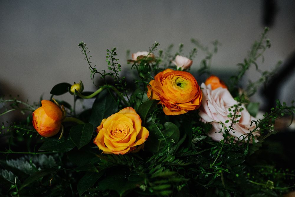 Romantic arrangement of orange ranunculus and yellow roses with delicate greenery in dim lighting.