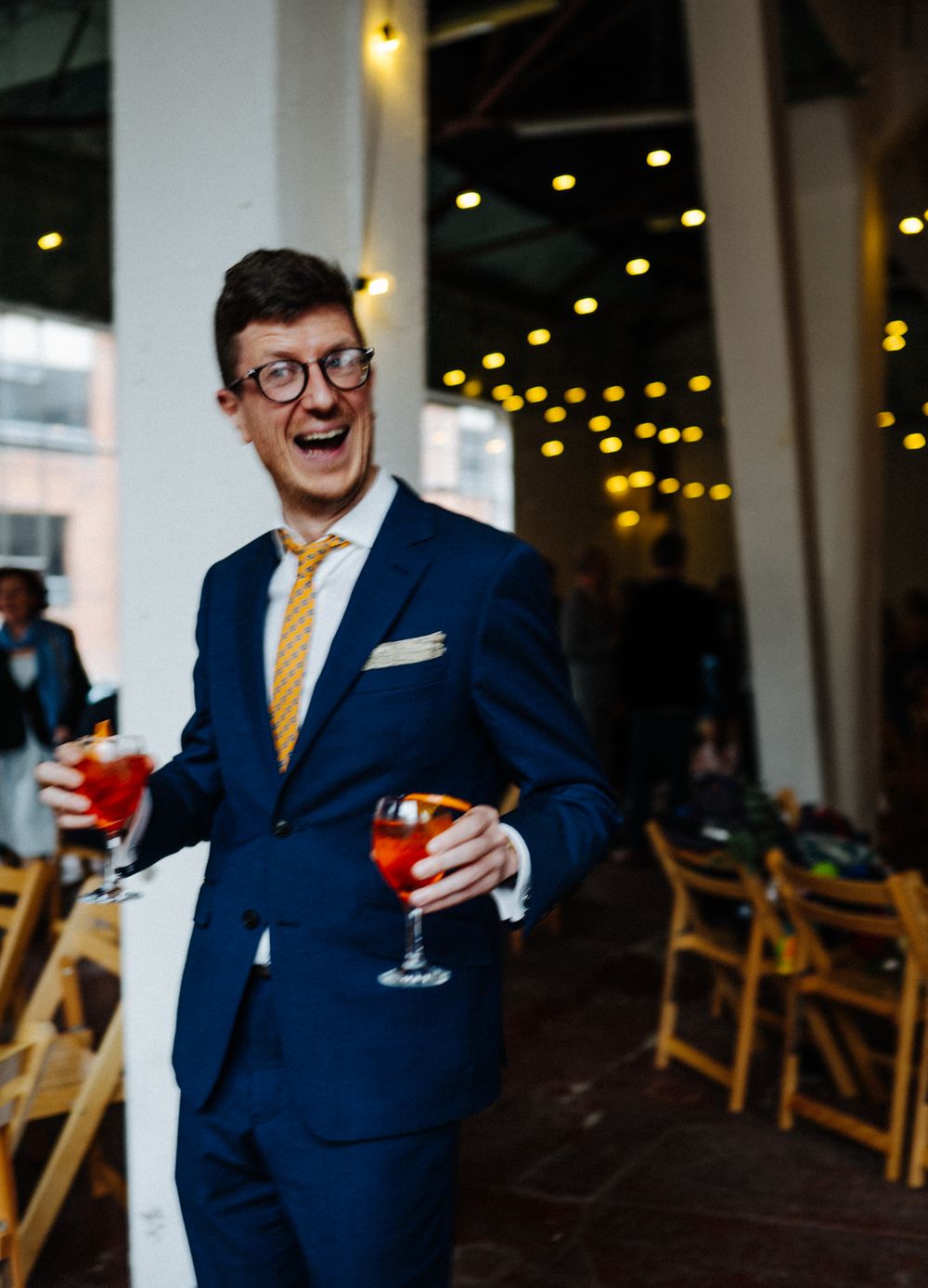 Wedding guest in blue suit and yellow tie laughing while holding cocktail.