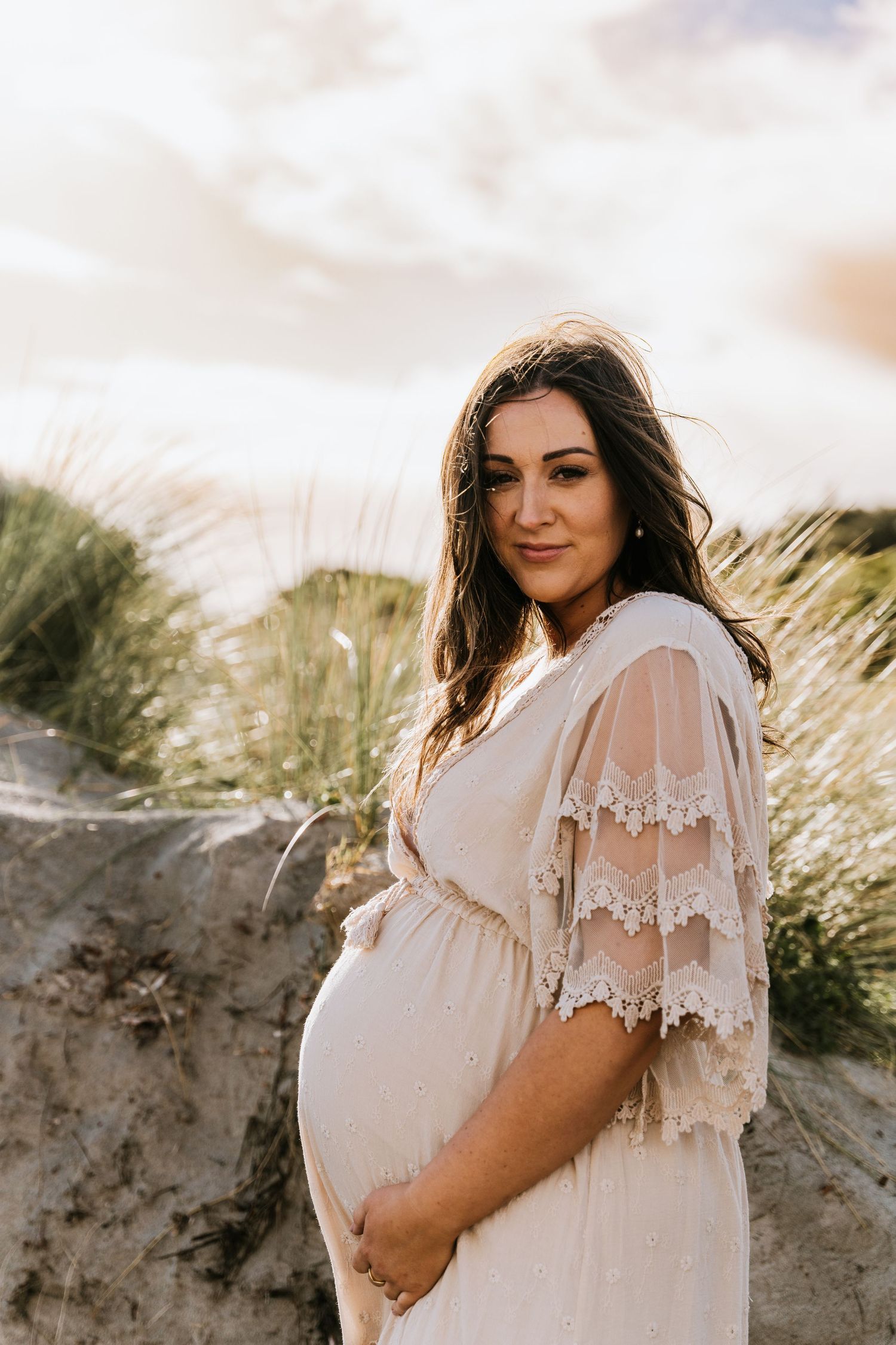 Expectant mother in lace dress poses against sand dunes during golden hour photoshoot.