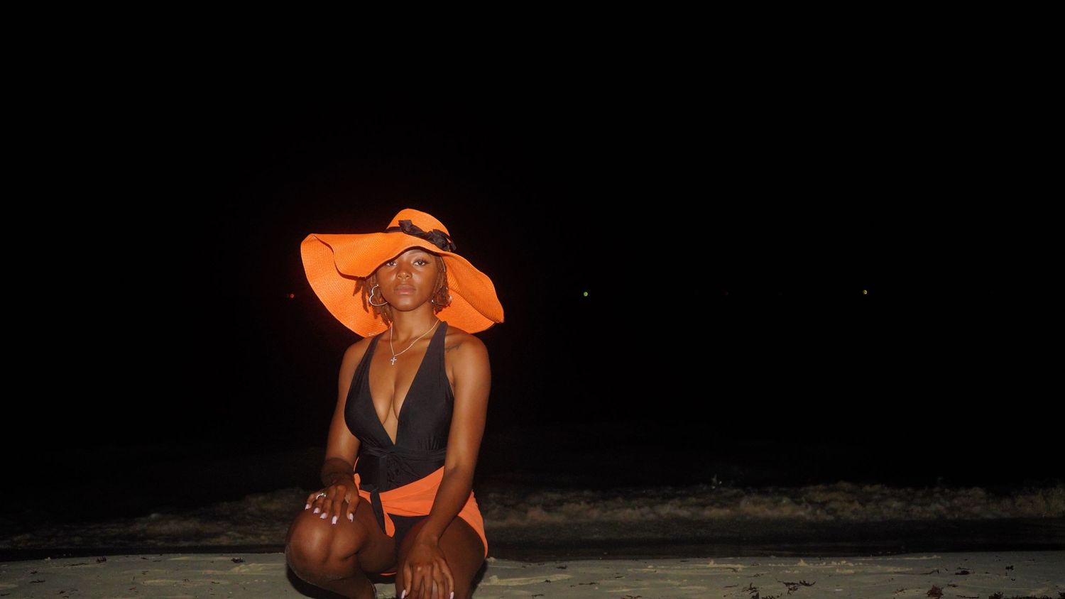 Figure in orange beachwear and sun hat poses against dark background at night.