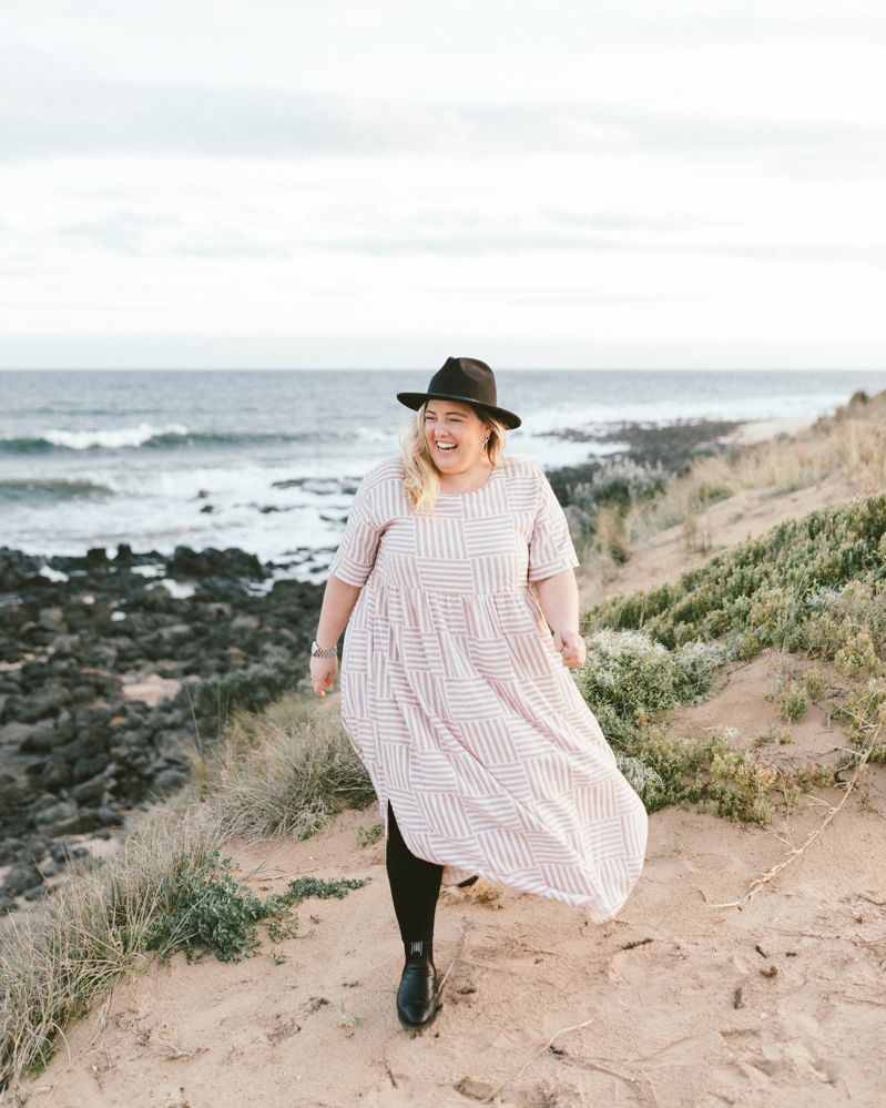 Woman in flowing dress and hat walks along coastal path with ocean views.