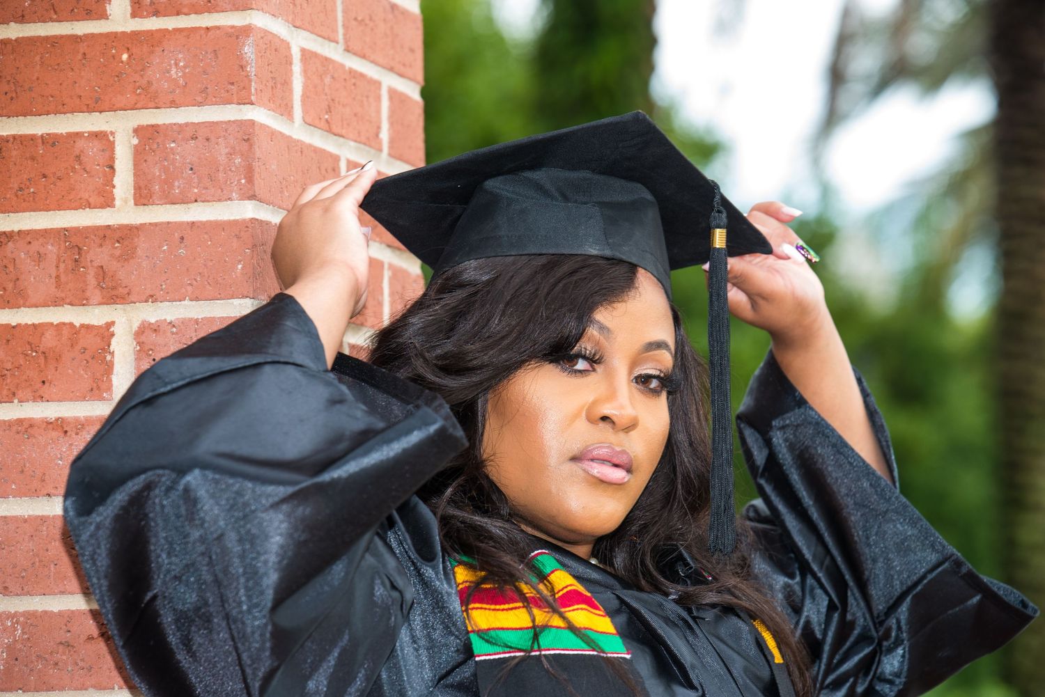 Graduate adjusts black cap with colorful stole against brick background.