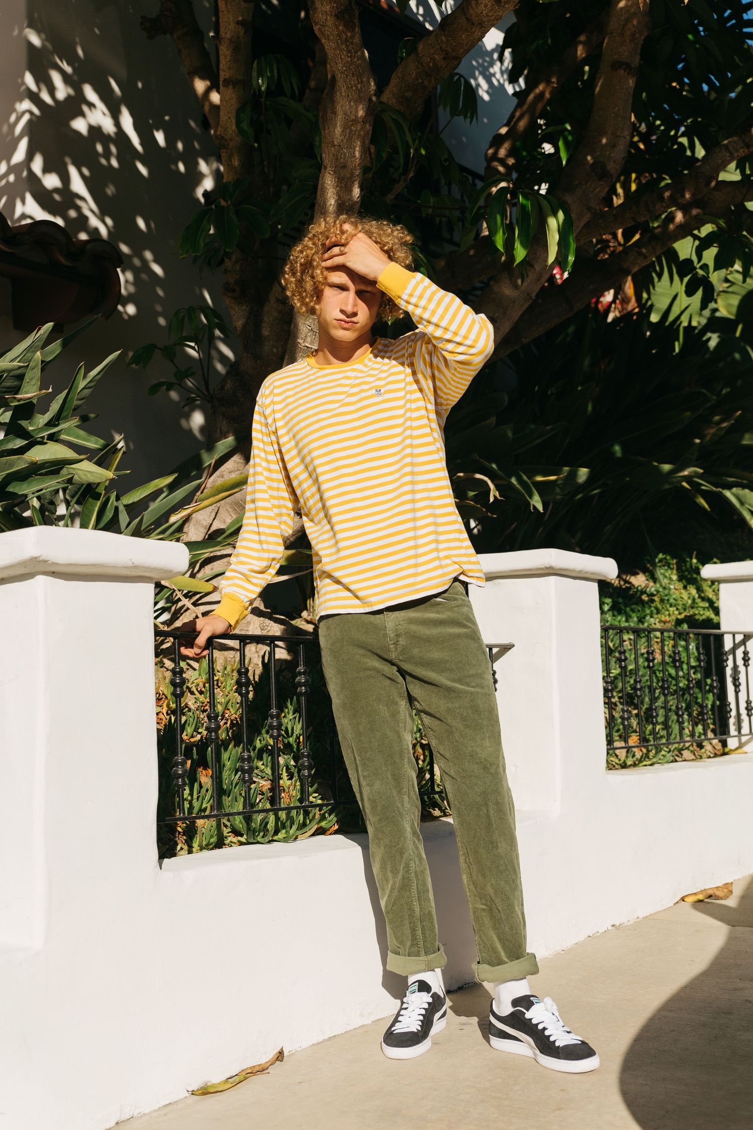 Curly blonde male model wearing yellow striped tee and olive corduroy pants in direct sunlight, San Clemente beach fashion editorial