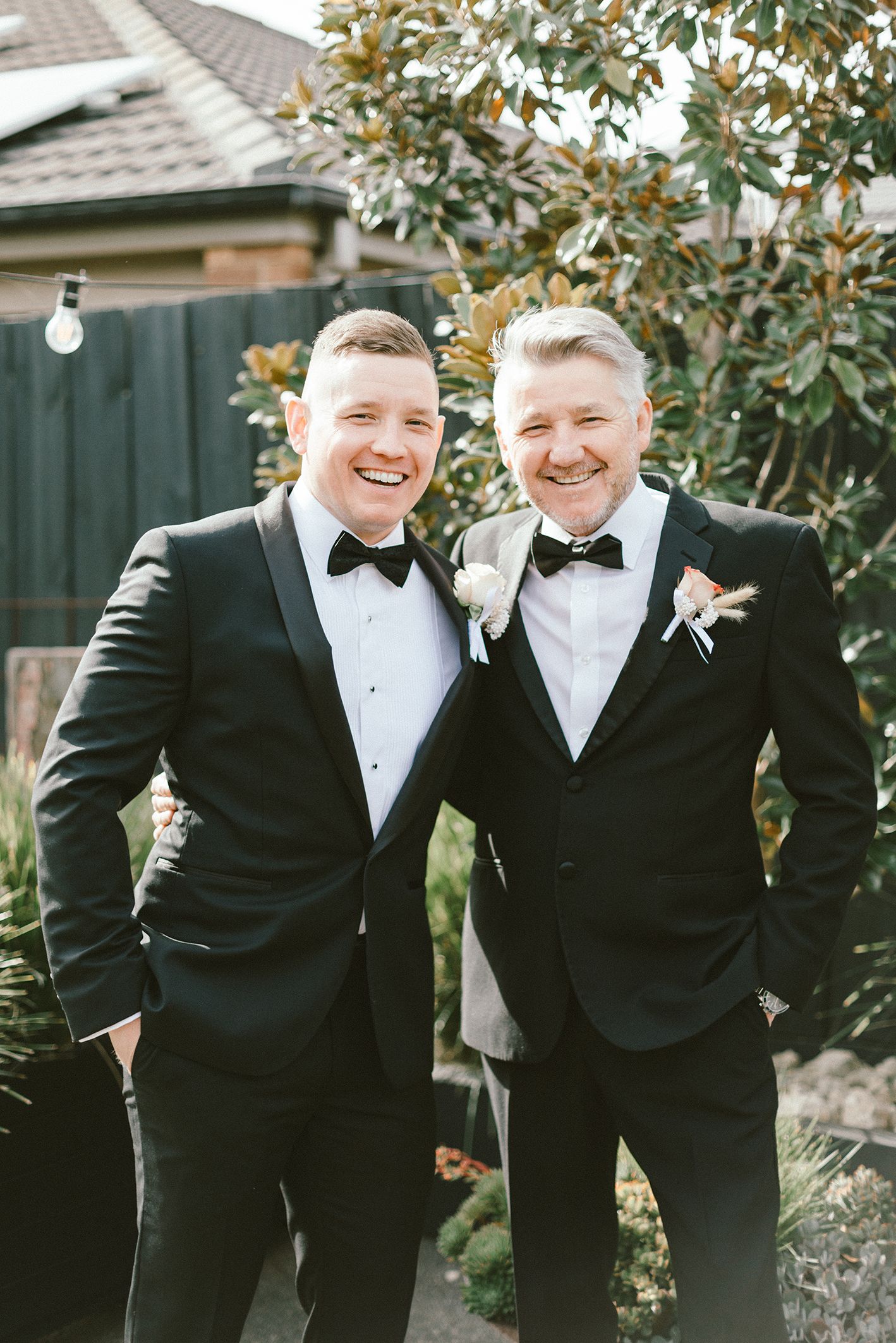 Two people in black tuxedos share a joyful moment in front of a magnolia tree.