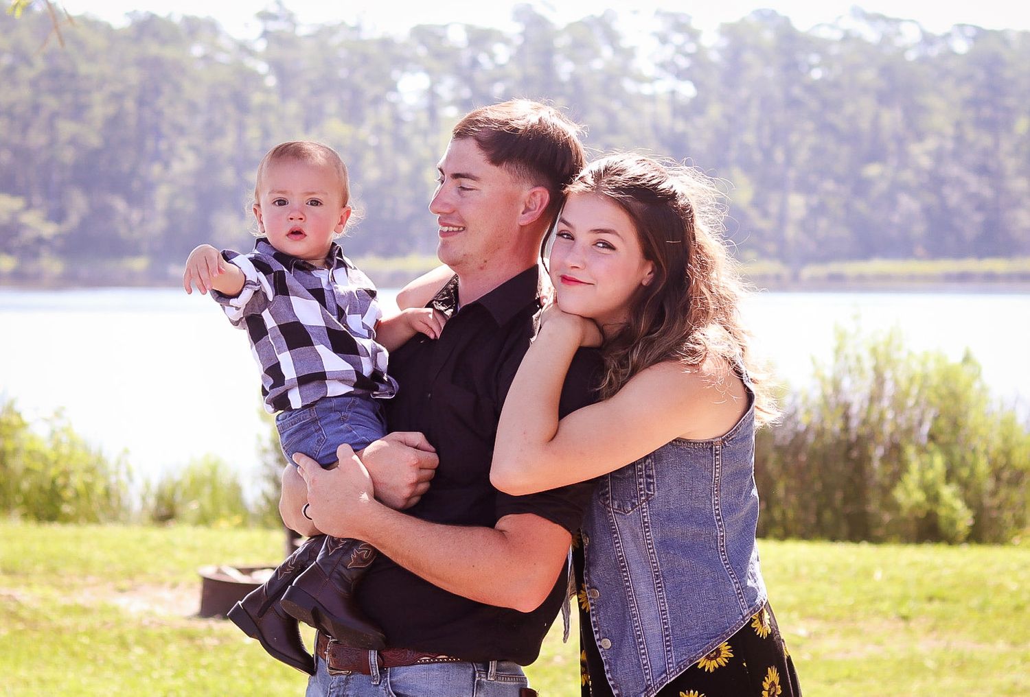 A couple smiles outdoors near a lake while holding their baby boy in a plaid shirt.