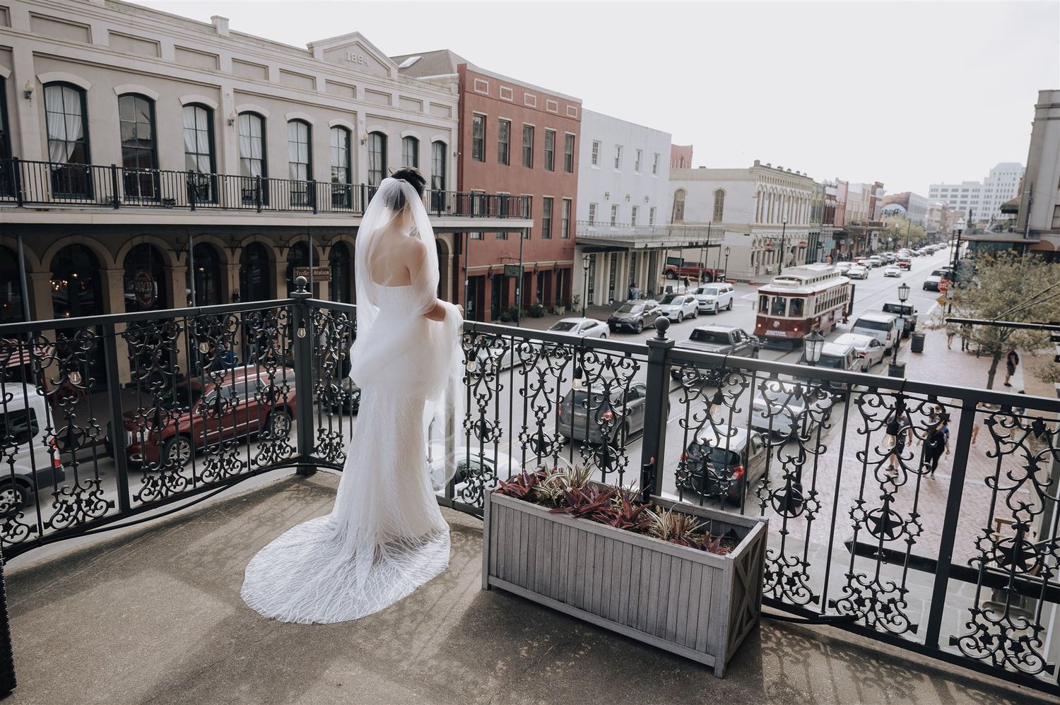 A bride in a white wedding dress stands on an ornate balcony overlooking a historic city street.