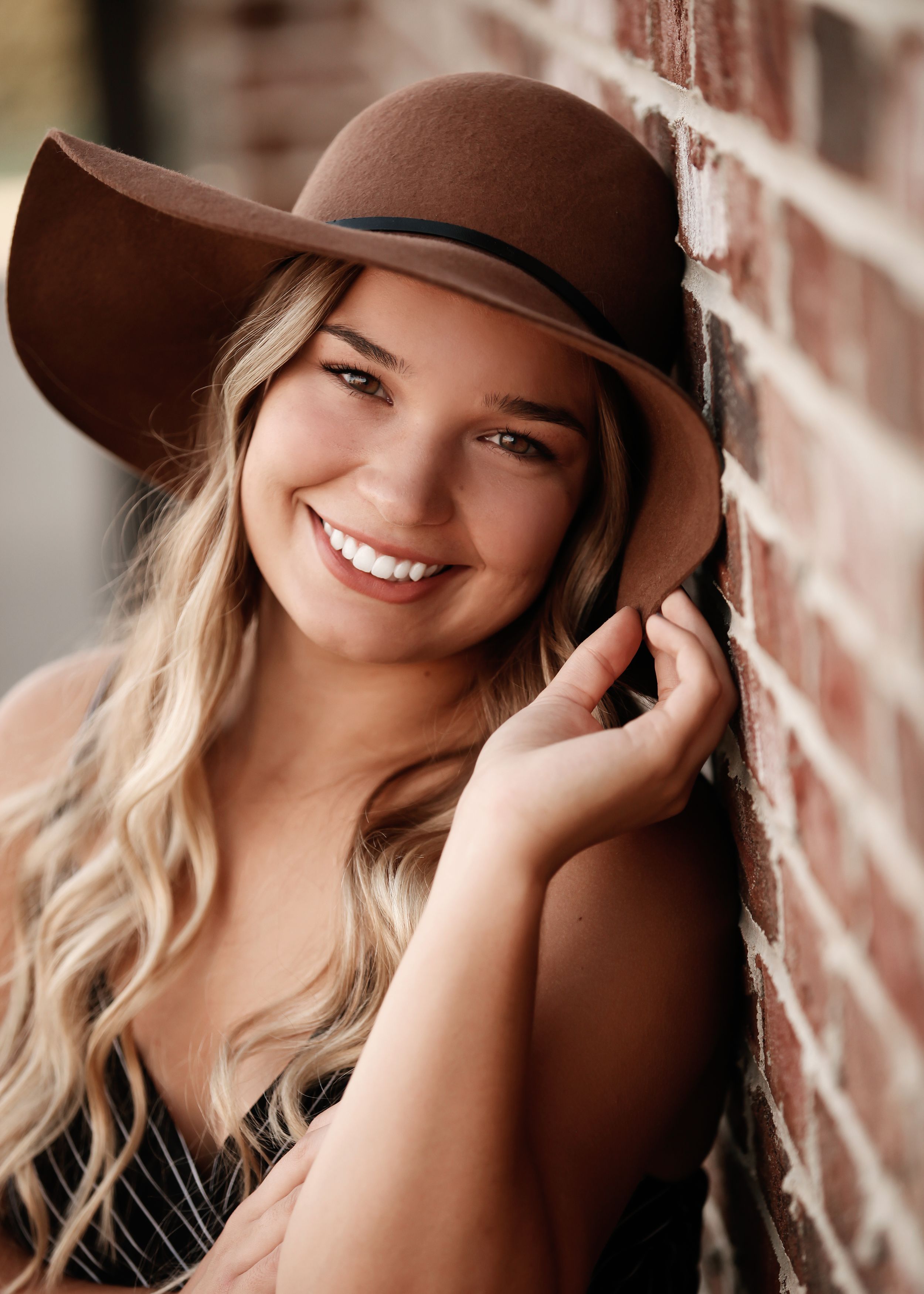A brown wide-brimmed hat complements a casual pose against a rustic brick wall.