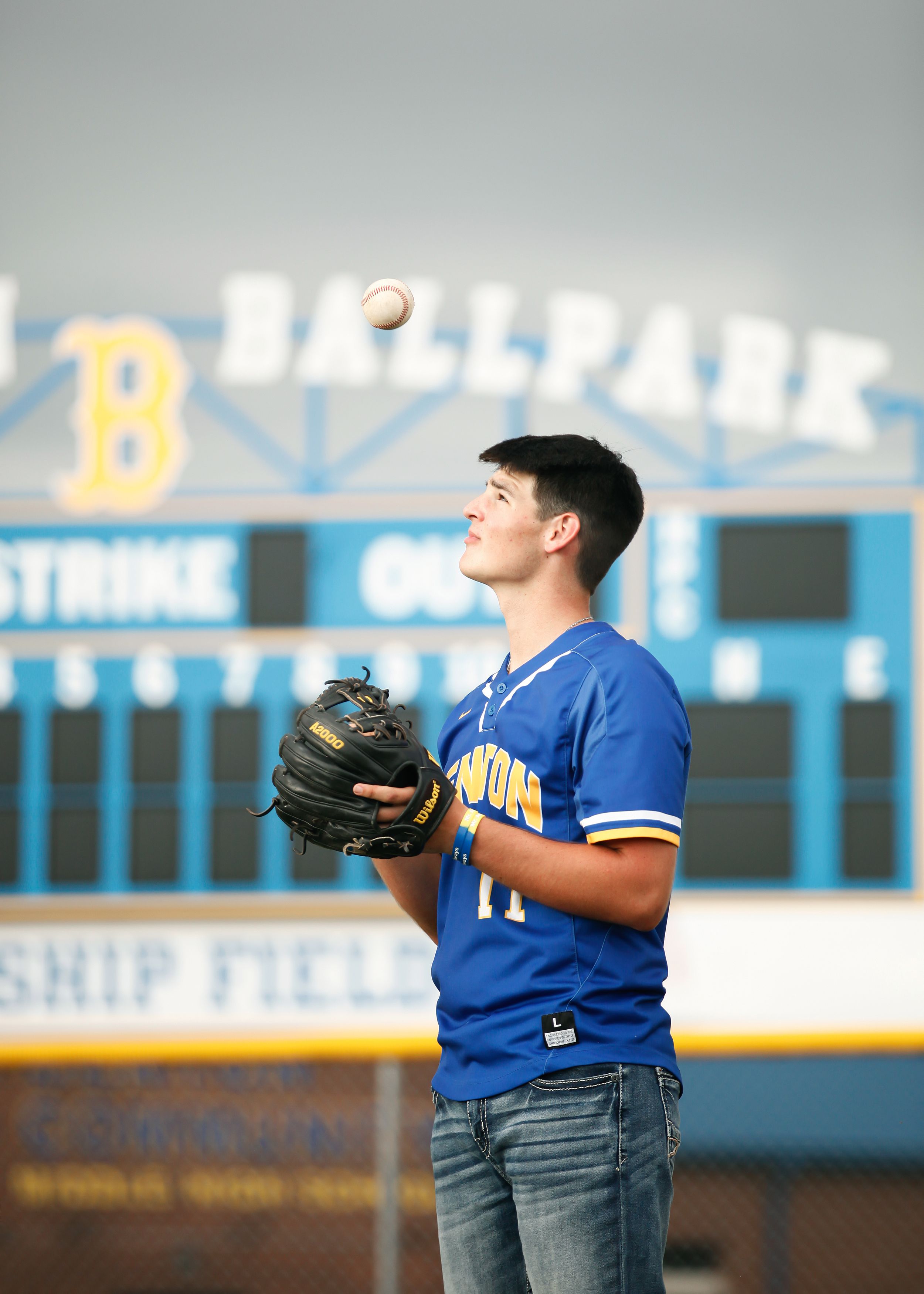 Baseball player in blue jersey practices catching and throwing on field with scoreboard in background.