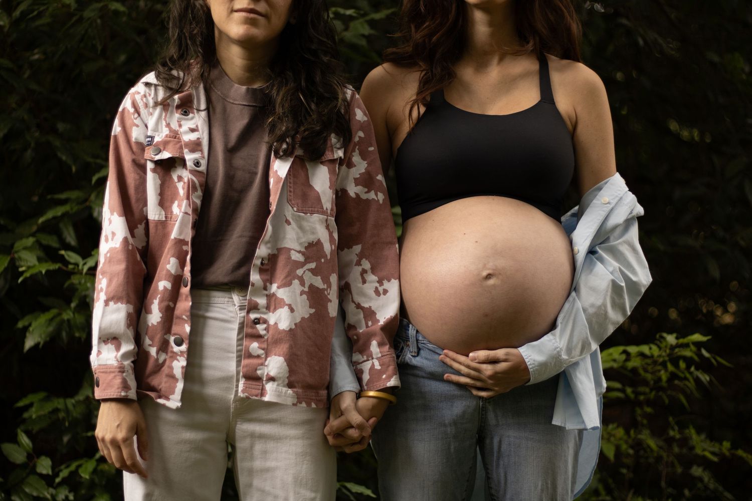Two individuals stand hand-in-hand outdoors, one in a pink jacket, the other in a black top, celebrating companionship and pregnancy.