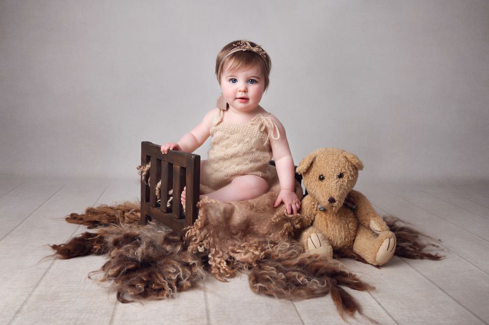 Baby sits with plush toy dog and rustic wooden props on light background for portrait session.